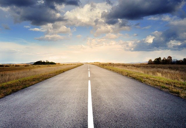 Empty countryroad in flat countryside, some trees in the far distance, cloudy sky